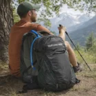 Man outdoors with hiking gear, Camelbak Magnus Hydration Pack, trekking pole, and mountain view.