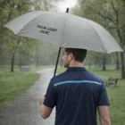Man holding Averie Umbrellas - Elite, gray with 'Your Logo Here', on wet park path.