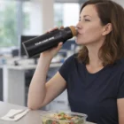 Woman drinks from a Bari Vacuum Bottle with a logo at a table with a salad.