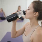 Woman in workout clothes drinks from Kamilah Leakproof Glass Vacuum Bottle labeled YOUR LOGO HERE.