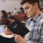 Young man writing in a Moleskine Pro Hard Cover Notebook with Your Logo, seated in a lecture hall.
