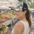 A woman in a Bryce Custom Mesh Cap smiles at an outdoor market with produce stalls behind her.