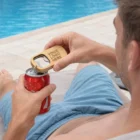 Man by a pool opens a soda can with a Bolsover Bottle Opener Key Ring labeled Your Logo Here.