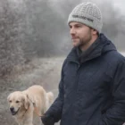 Man in winter coat and Promotional Cable Knit Beanie walks a golden retriever on a frosty path.