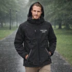 Man in a black Sols Rock Mens Softshell Jacket with Your Logo Here, standing on a park path.