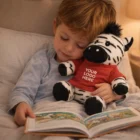 Young boy reading in bed, hugging a Zebra Branded Plush Toy in a red Your Logo Here shirt.