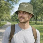 Man wears a green Clovelly Logo Branded Bucket Hat and gray shirt in a tree-filled park.
