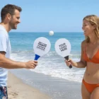 A man and woman play Play Now! Paddle Ball Games on the beach with branded paddles, smiling.
