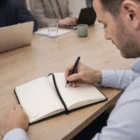 Man writing in a Pierre Cardin Biarritz Notebook Gift Set at a meeting table with laptops and cups.