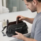 Man puts a bottle in a Pierre Cardin Leather Toiletry Bag on a bathroom counter.