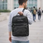 Man carrying a Pierre Cardin Leather Backpack with YOUR LOGO HERE, walking on a paved path.