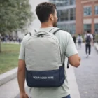 Man on a campus path wearing an Essex Laptop Backpack with "YOUR LOGO HERE" on the front.