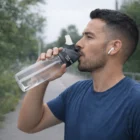 Man drinks from Oshawa Customised Bottles outdoors, wearing wireless earbuds.