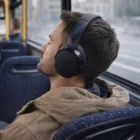 Man wearing Skullcandy Hesh Evo Headphones relaxes on a bus, eyes closed, in a brown jacket.