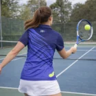 Woman in Ladies Sublimated Polos plays tennis, preparing to hit a ball on an outdoor court.