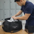 Man in gym clothes packs an Aspen 20L Duffle Bag labeled YOUR LOGO HERE in a locker room.