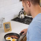 Man cooking bacon and eggs beside Ardmore Bamboo Weather Stations showing time and temperature.