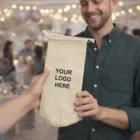 Smiling man receives a Madeira Cotton Bottle Gift Bag with "Your Logo Here" at a festive event.