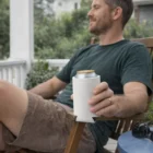 Man relaxes on a porch with a canned drink in a Prairie Stubby Cooler, wearing casual clothes.