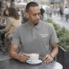Jude relaxes outdoors in Jude Mens Polos, holding a coffee cup with Your Logo Here.
