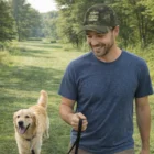 Man in blue shirt wears Nalem Camouflage Cap while walking golden retriever in grassy park.
