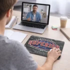 Man video chatting on laptop at desk with coffee, notebook, and Ellicott Mouse Mats.