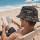 Man reading on a Hobart beach, wearing Hobart Camouflage Bucket Hats with YOUR LOGO HERE.