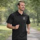 Man jogging in a Men Ace Performance Polo Shirt with Your Logo Here text on a park path.