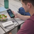 Man charging phone with Natura Wheatstraw Power Bank at outdoor table, salad and water nearby.