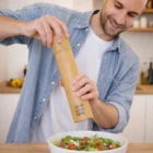 Man in blue shirt seasoning salad with Rook Bamboo Pepper Mills, smiling in his kitchen.