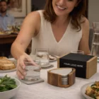 Smiling woman sets glass on a Keepsake Pebble Coaster Set at a dining table with matching coasters.