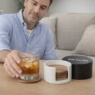 Man placing iced drink on Keepsake Coaster Set beside coaster holder on table.
