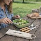Woman at picnic table uses Reusable Stainless Steel Cutlery Set in branded fabric pouch.