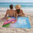 A couple sits on the sand with a Dune Poly Cotton Beach Towel showing “SUMMER Time” and a pink flamingo.