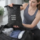 Woman holding a black Hilliard Nylon Lightweight Bag above packed clothes and shoes.