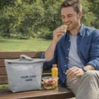 Man eating sandwich on bench with Naples Insulated Lunch Bag, fruit salad, and juice on table.