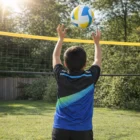 A boy in a Kids' Singlets sets a volleyball over an outdoor net on a sunny day.