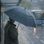 Person holding a Sumbrell Pongee Canopy Sports Umbrella at a bus stop in heavy rain as a bus nears.