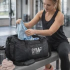 Packing gym clothes into an Urbangear Polyester Duffle Bag labeled GYM while on a bench.