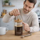 Smiling man using a Glass Coffee Plunger to brew coffee on a kitchen counter.