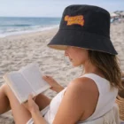 Woman in a Reversible Bucket Hat reading by the ocean on a sandy beach.