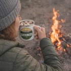 Person in a beanie drinks from 500Ml Enamel Mugs by a campfire outdoors.