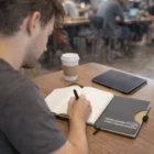 A man writes in a Thermo Pu Notebook at a table with coffee and a tablet nearby.