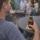 Man holding beer with Square Bottle Opener Coasters at outdoor table, barbecue and friends nearby.