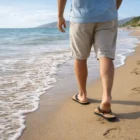 Man in shorts wearing Summer Fiji Flip Flops walking on the sandy beach shoreline.