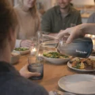 A hand pours water from the Dusk Carafe And Tumbler Sets at a dinner table with food and people.