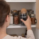 A woman places a Glass Onsen Canister with cork lid on a pantry shelf with spice jars.
