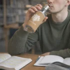 A person drinks from a Glass Bottles With Cork Sleeve while studying with open books at a table.