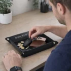A man puts keys, coins, phone, and wallet in a Leather Valet Tray on a wooden desk.