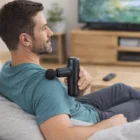 Man using a Handheld Massager on his shoulder while sitting on a couch at home.